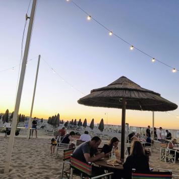 Beach at sunset with people dining under decorative lights.