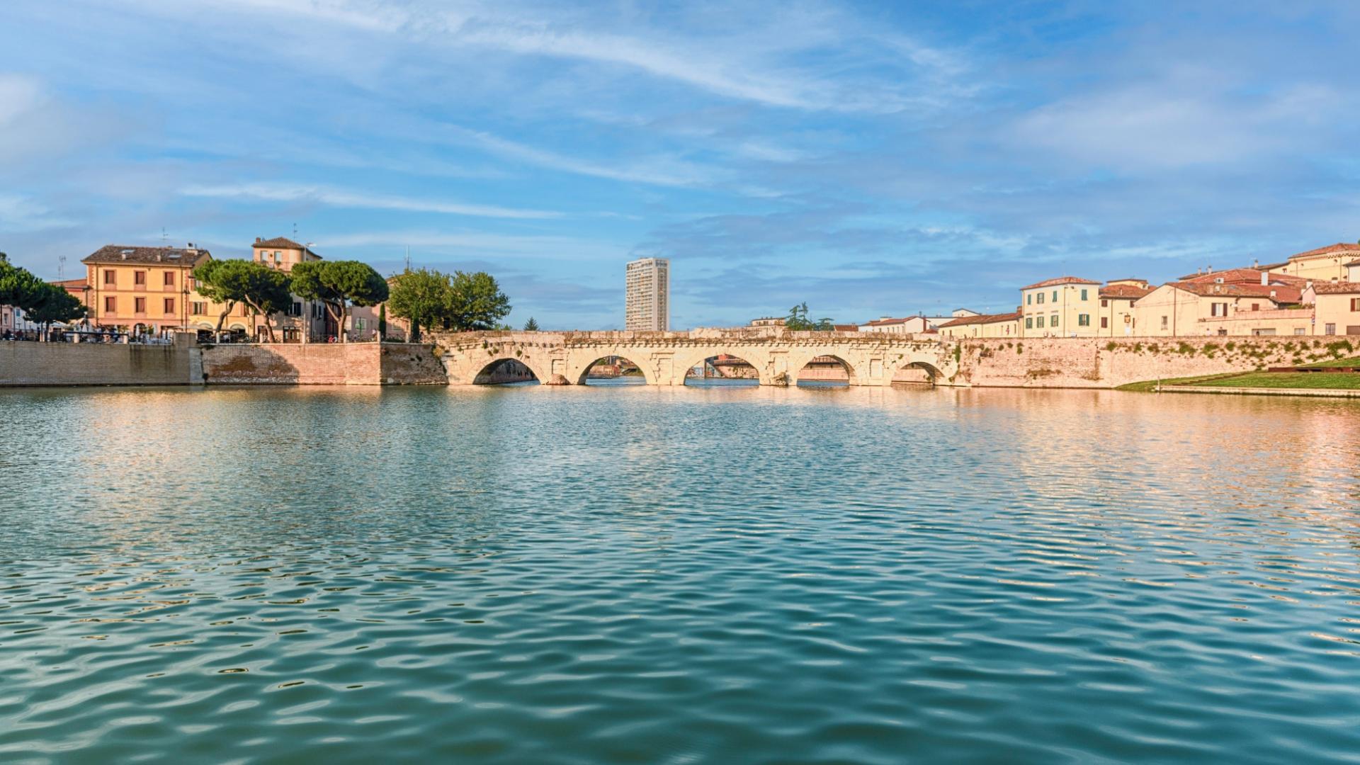 Ponte antico su fiume calmo, case colorate e cielo limpido, atmosfera serena e storica.