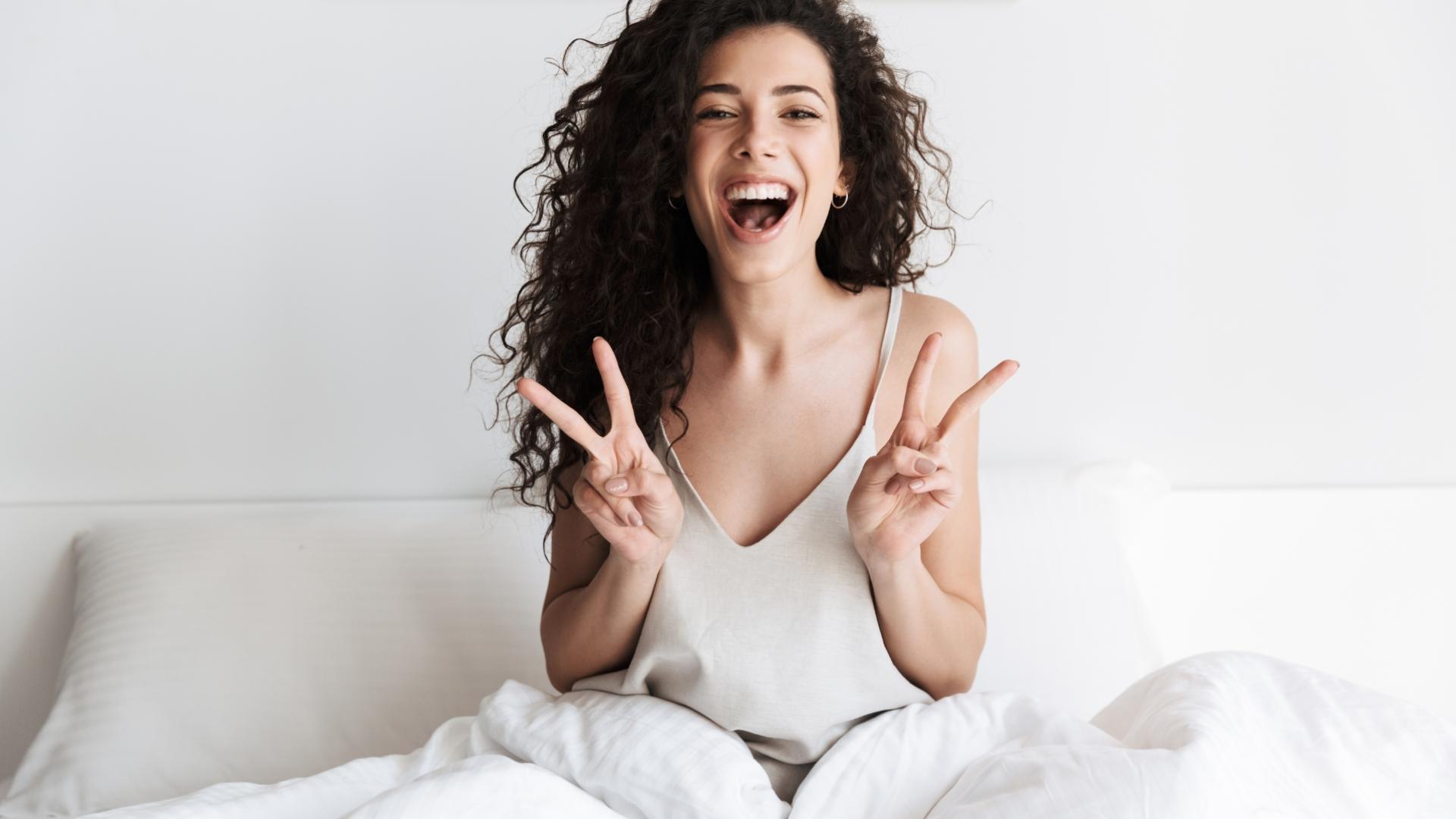 Donna sorridente a letto, capelli ricci, fa segno di pace con entrambe le mani.