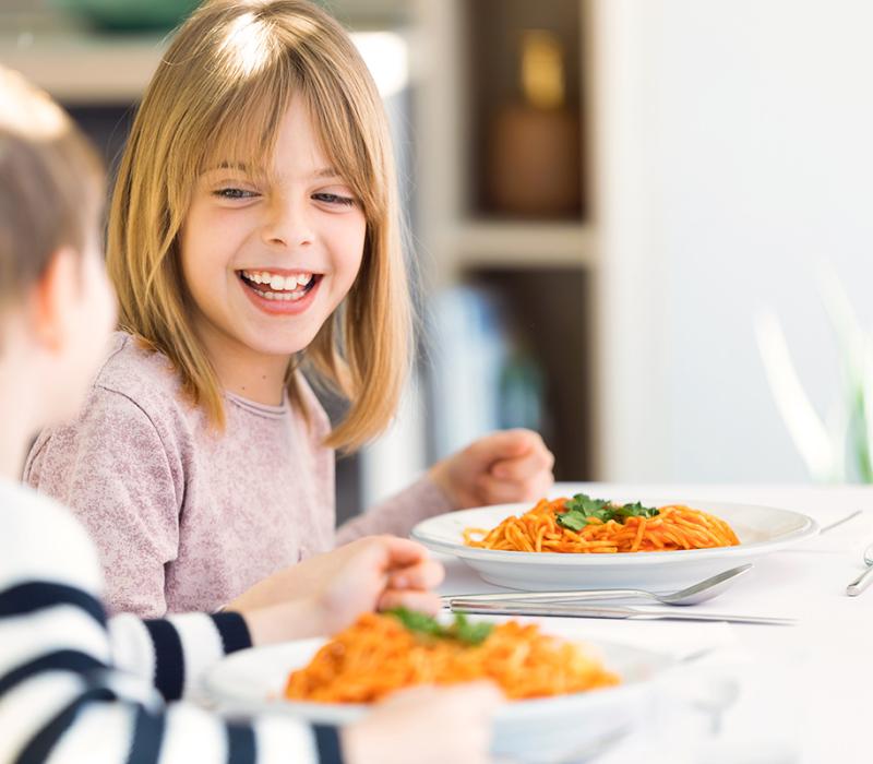Due bambini sorridenti mangiano spaghetti con salsa di pomodoro a un tavolo luminoso.